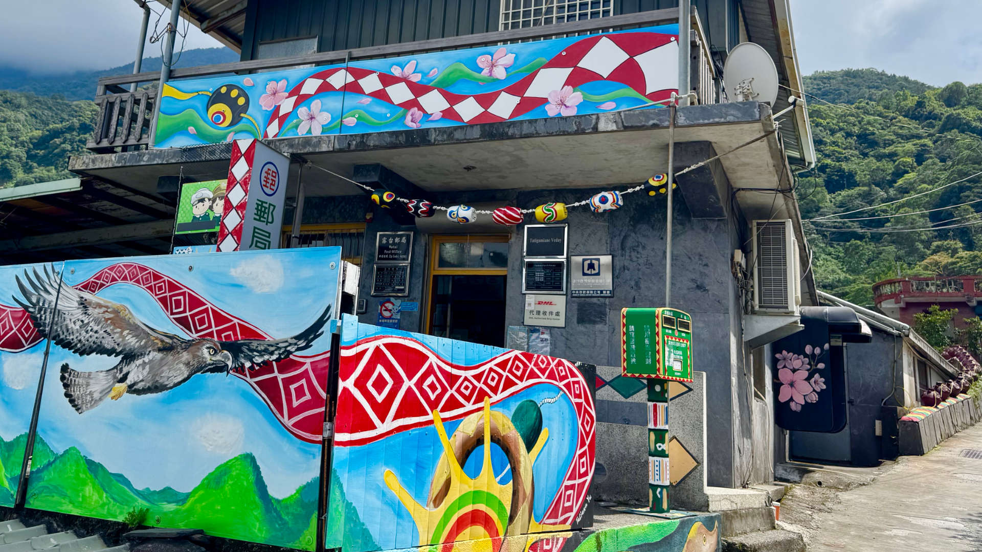 A compact two-story concrete building with vibrant artwork on the second-floor balcony and a fence out the front. Next to the entrance is a typical Taiwanese mailbox, but with additional colorful geometric patterns on it.