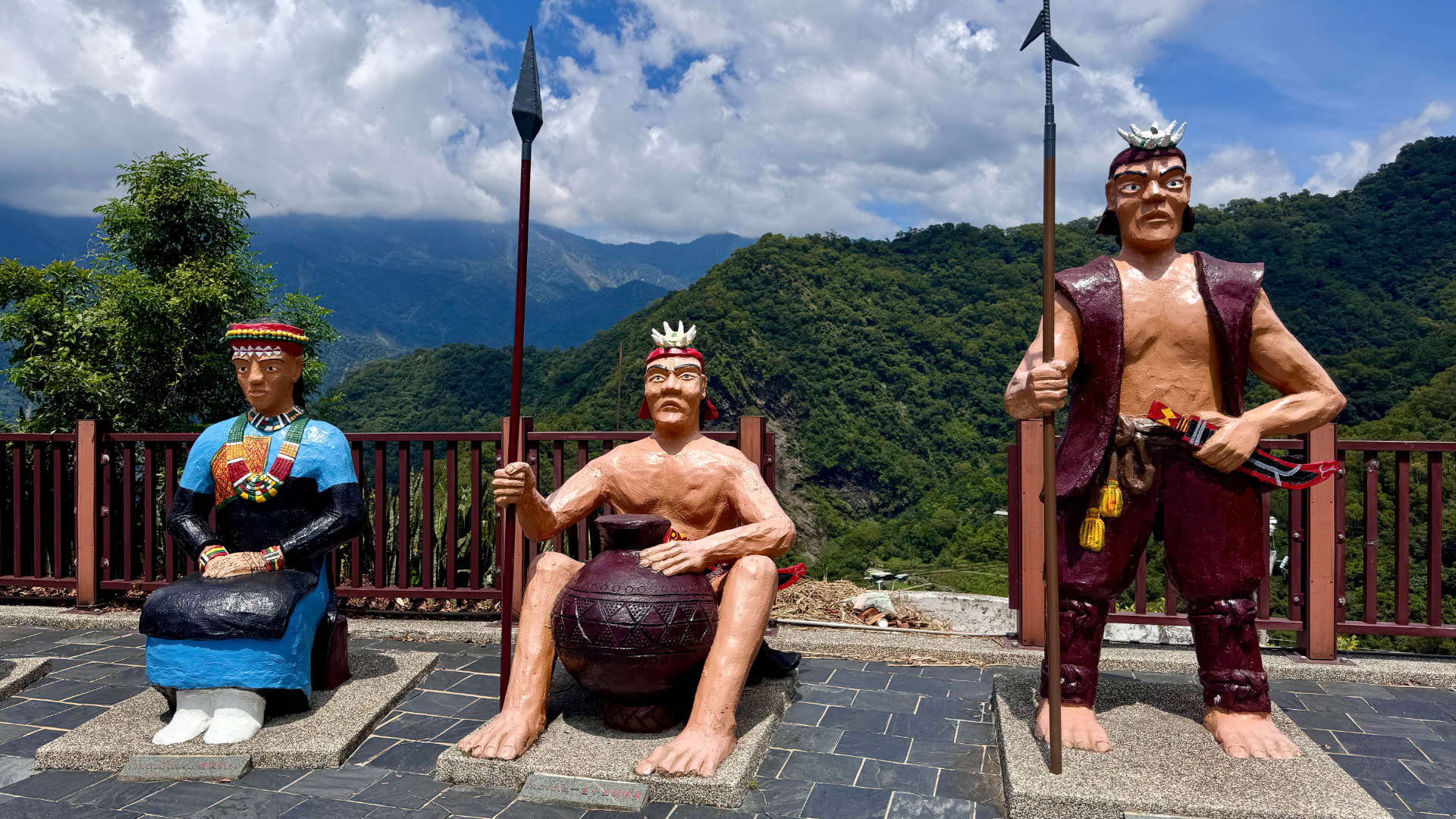 Three large Rukai tribal statues in Wutai Township, with a dramatic view of forest-covered mountains in the distance.