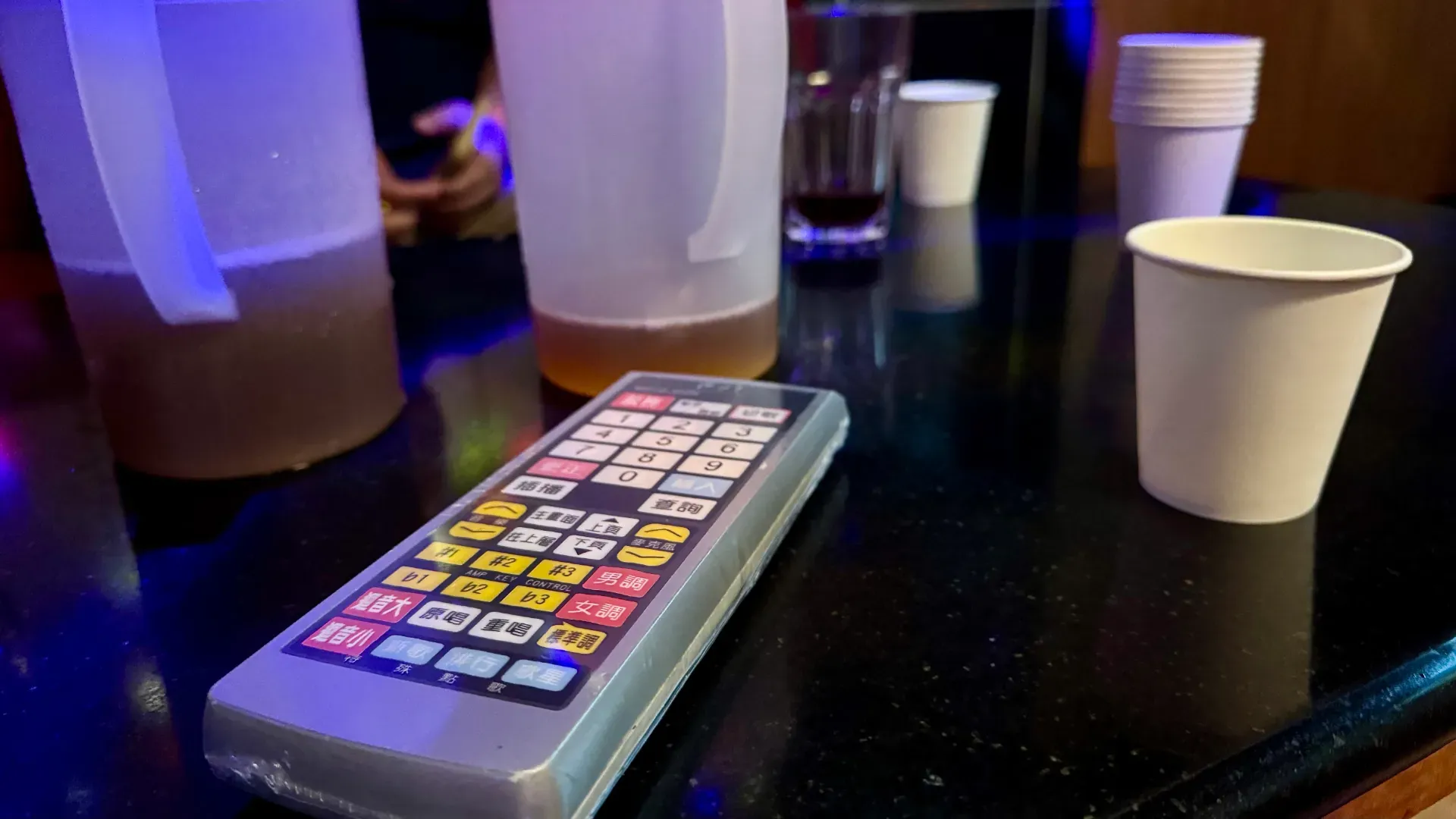 A complicated plastic-wrapped remote control on a coffee table amongst jugs of tea and paper cups.