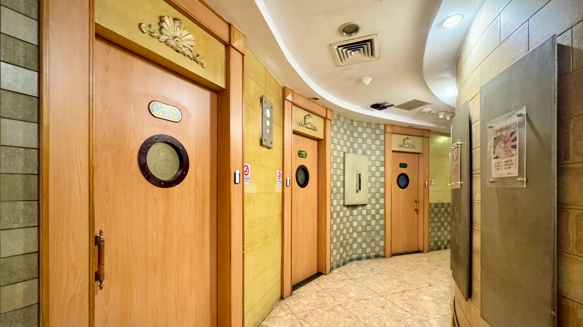 A curved corridor inside Soaring Spirit KTV (神采飛揚KTV) in northern Kaohsiung. There are a series of wooden doors labelled A-21 to A-23, each of which has a porthole window at eye height.