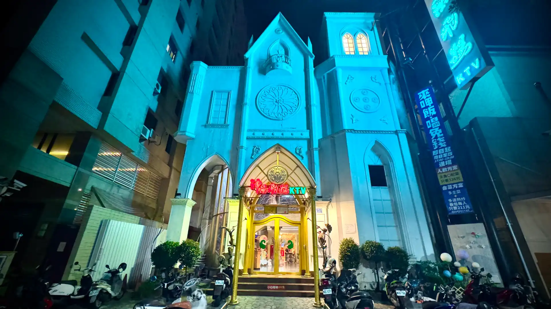 The exterior of Soaring Spirit KTV (神采飛揚KTV) in northern Kaohsiung. The building looks like a two-story white church with a three-story belltower. Many scooters are parked outside.