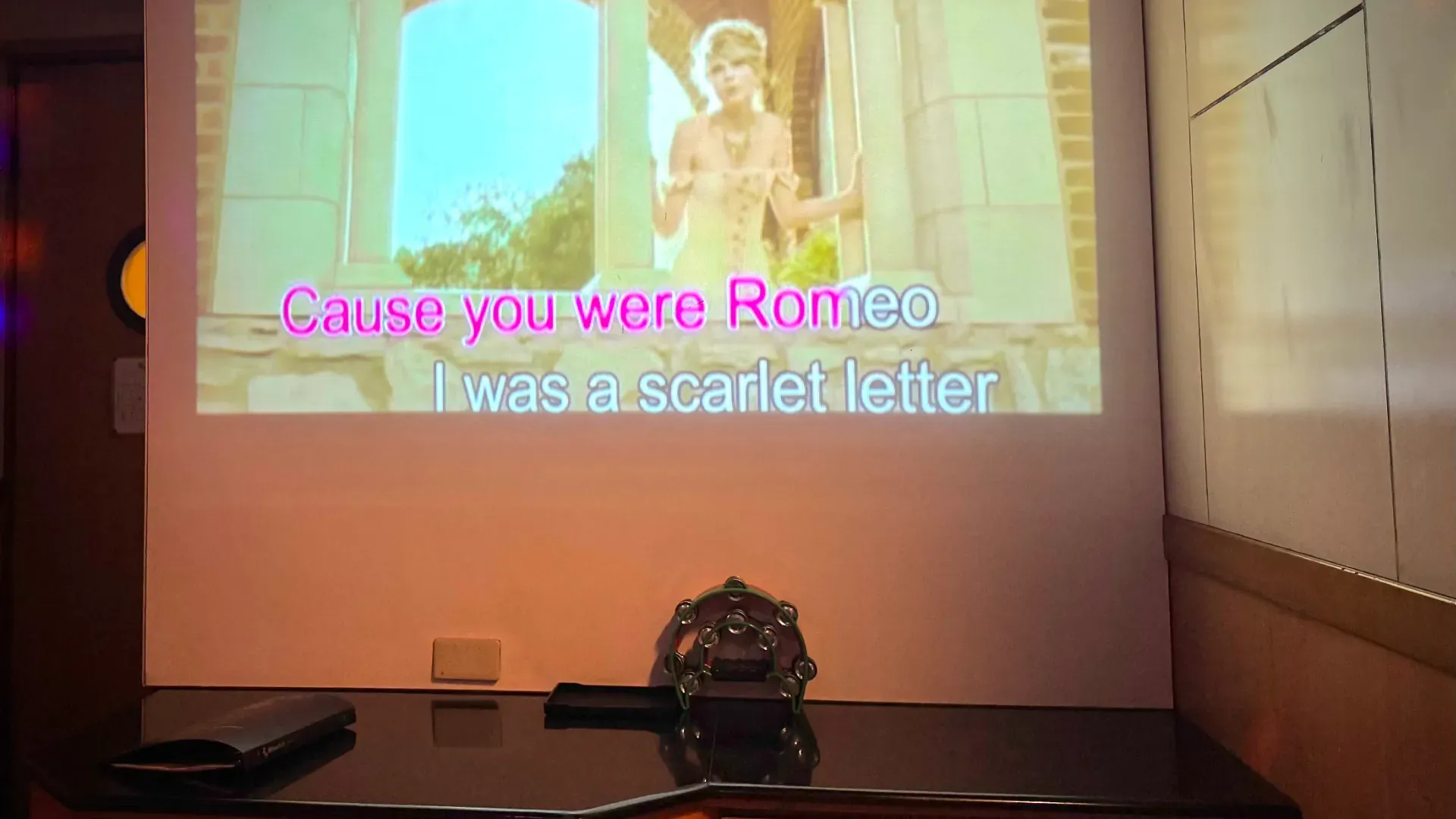 Taylor Swift’s music video for ‘Love Story’ projected onto a wall in a private KTV room in Taiwan. Below the video, a pair of tambourines sit unused on a coffee table.
