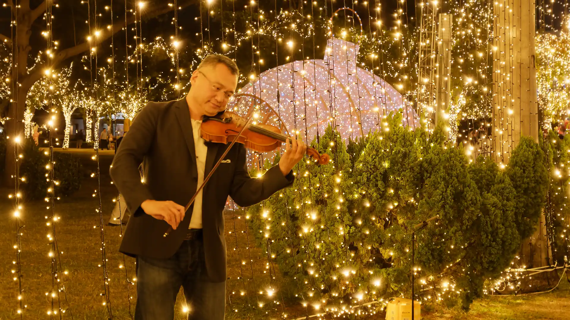 A smartly-dressed middle-aged busker playing a violin in front of Christmas lights.