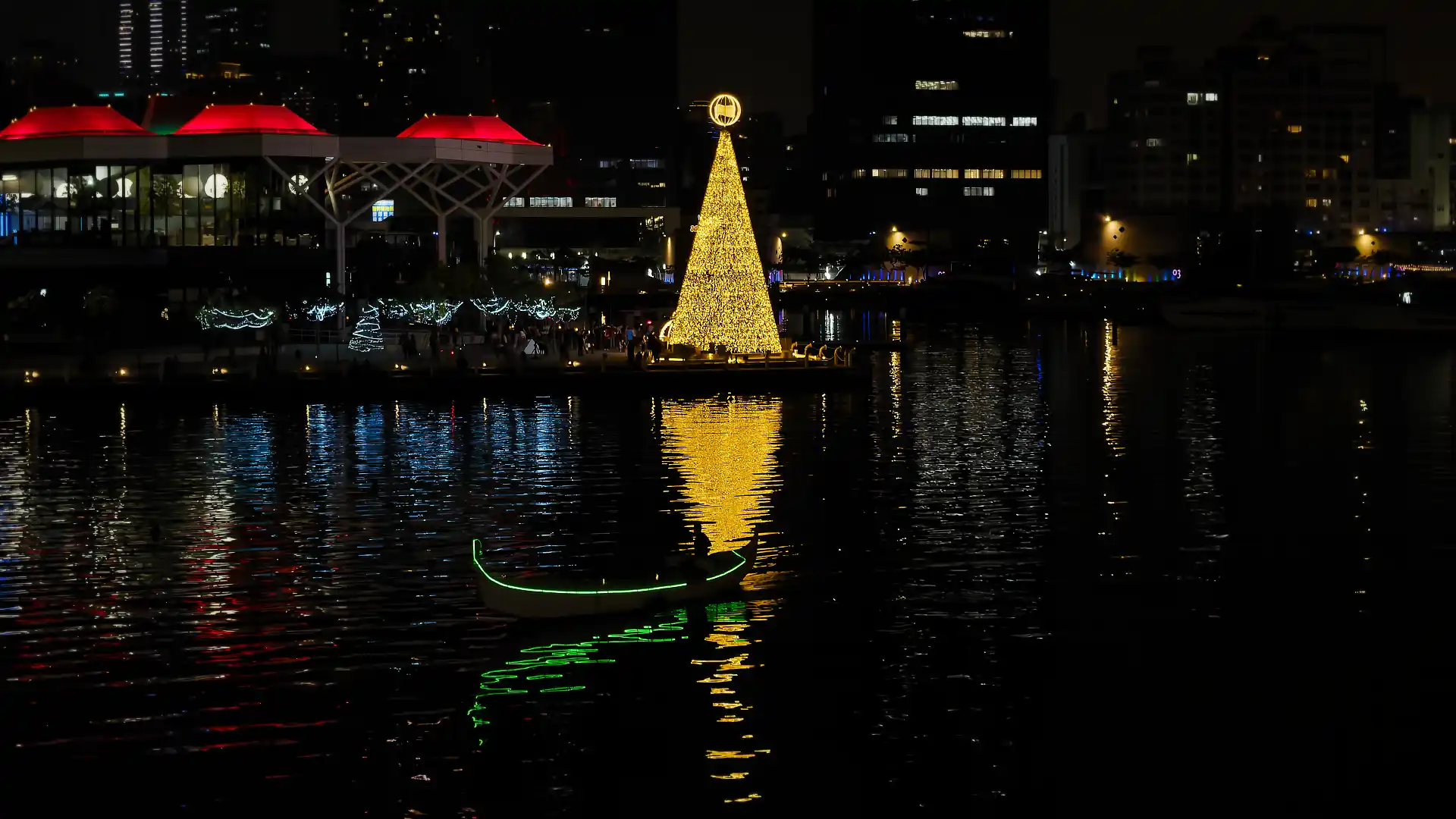 A Venetian-style gondola passing on the water in front of a golden Christmas tree at Kaoshiung harbor. The gondola driver is standing, and silhouetted against the Christmas tree's reflection.