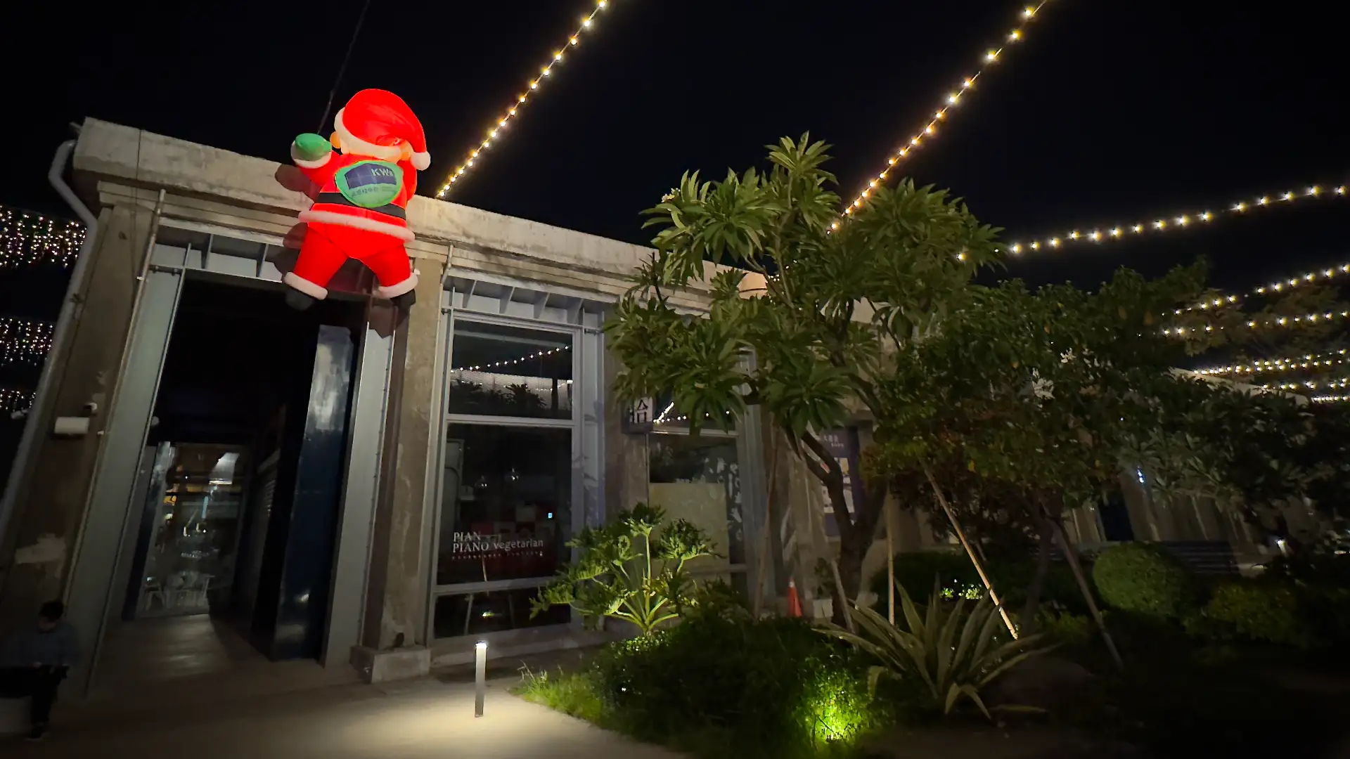 Festoon lights hang over a garden at Pier 2 Art Center. A large inflated Santa appears to be scaling the wall.