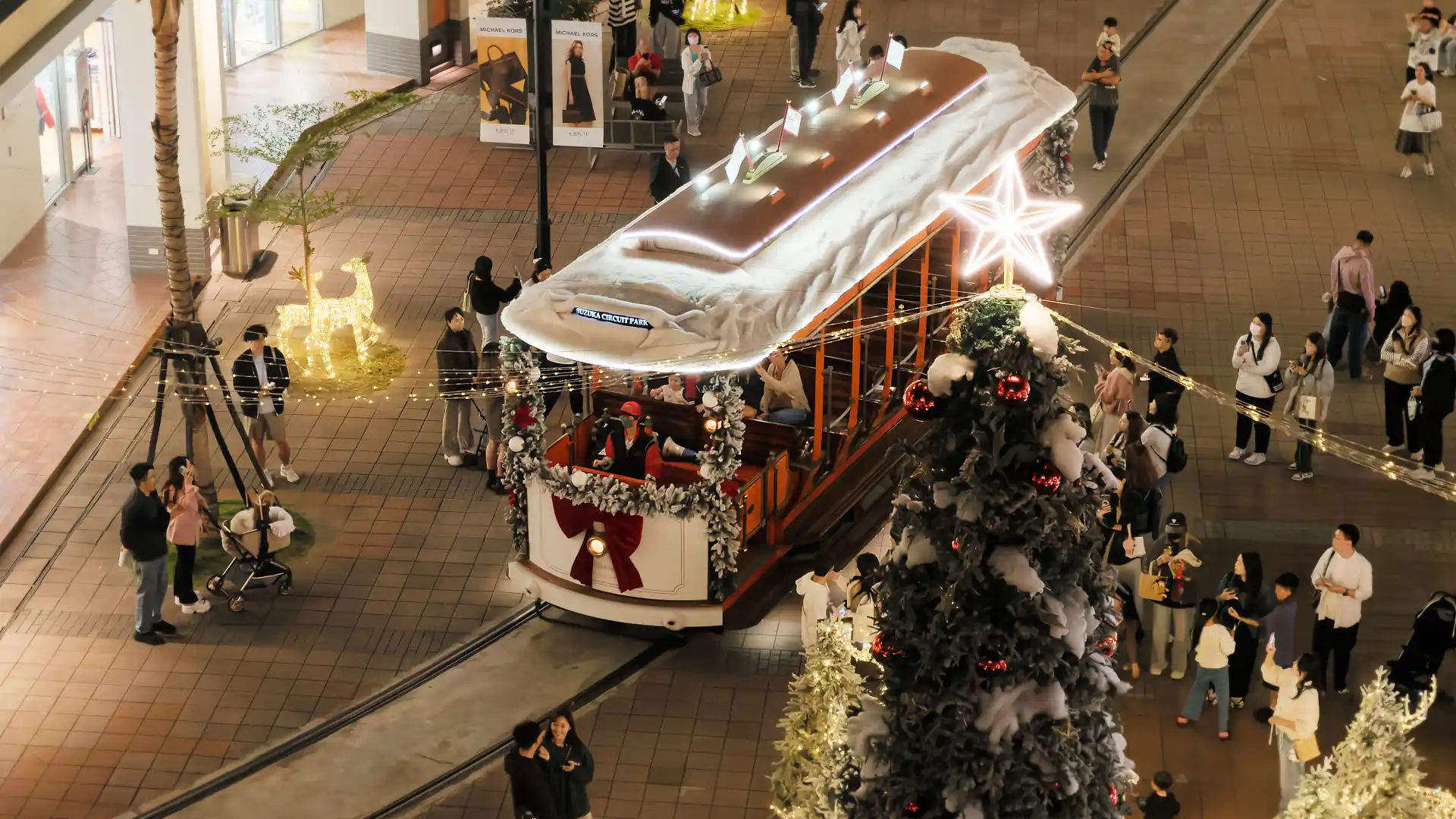 Close-up of a tram moving through SKM Park shopping mall's outdoor square. It is decorated in lights and fake snow. It is passing a large Christmas tree.