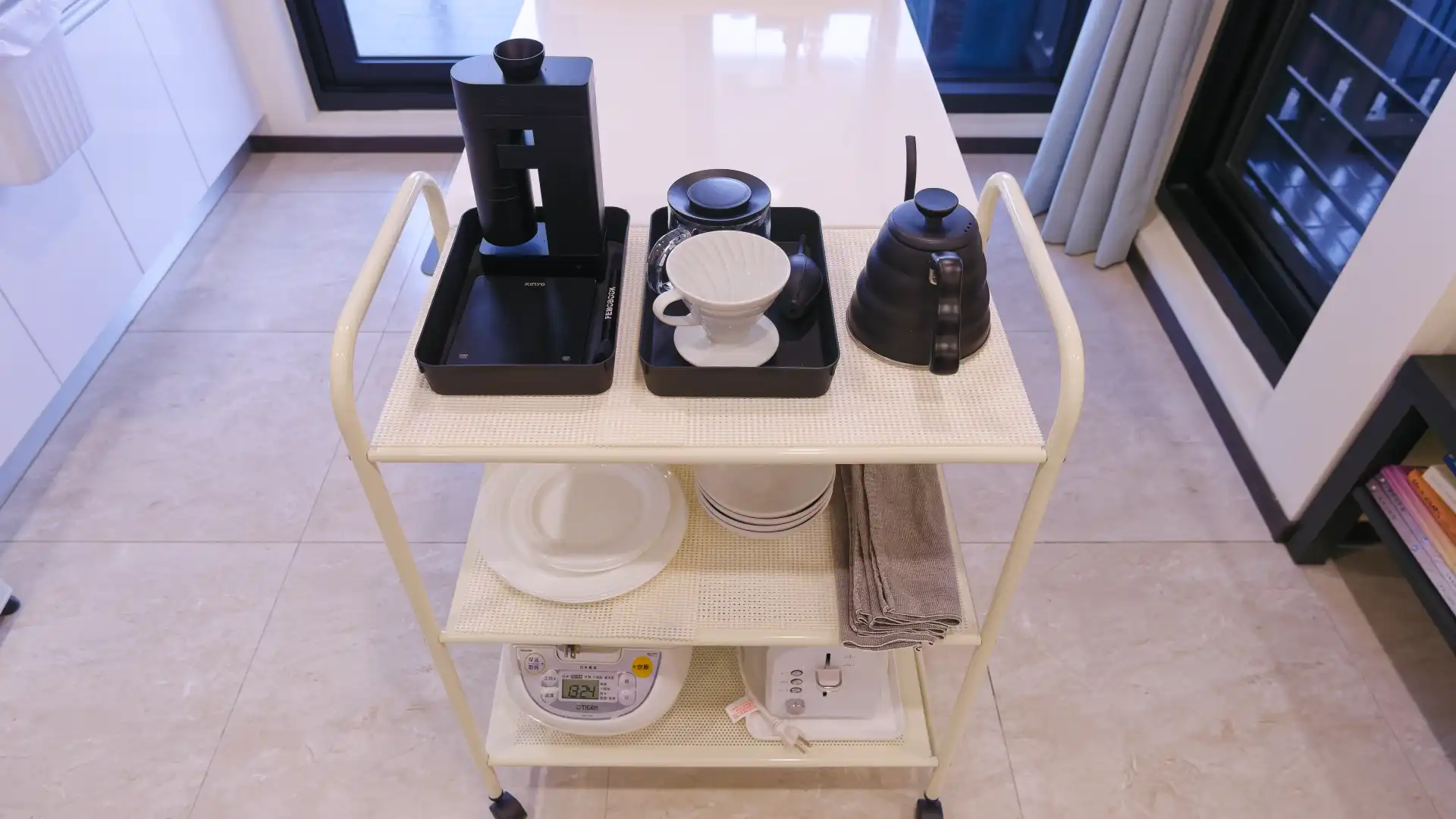 A kitchen trolley with three shelves, containing appliances and plates and coffee-making equipment.