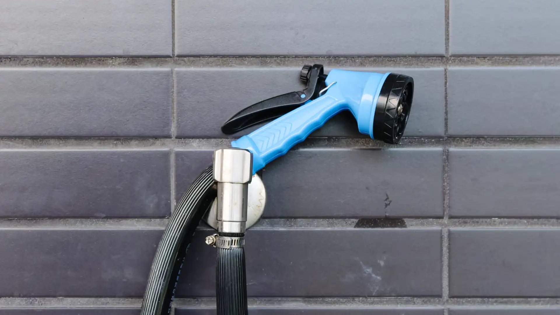 A dripping garden spray gun, attached to a hose, and nestled on top of a wall-mounted outdoor garden tap.
