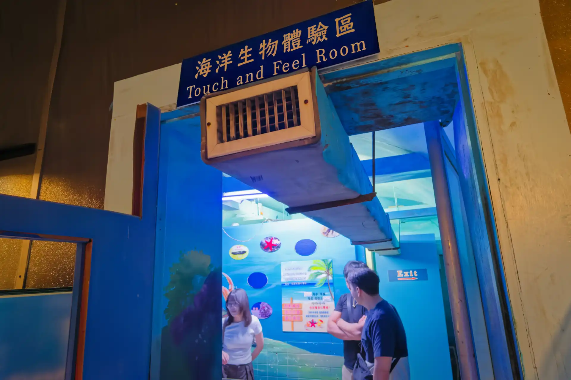 Three people standing in an underground concrete-lined room with the sign “Touch and Feel Room” above the doorway.
