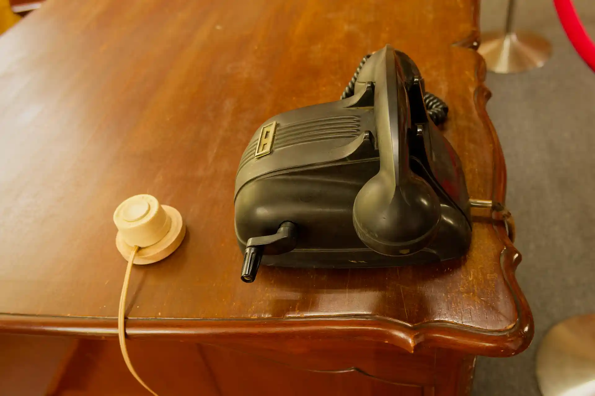 A black telephone on a desk. It does not have a rotary dial or any buttons. It has a hand crank on the right-hand side. There is also a white plastic button affixed to the top of the desk.