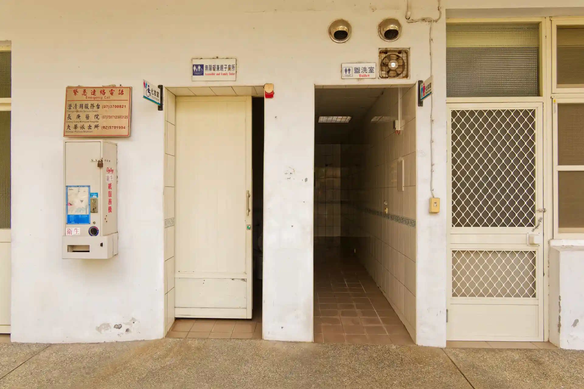 A public toilet with two entrances, one accessible and one all-gender. Next to the accessible entrance is a wall-mounted vending machine with faded stickers and signage on the front.