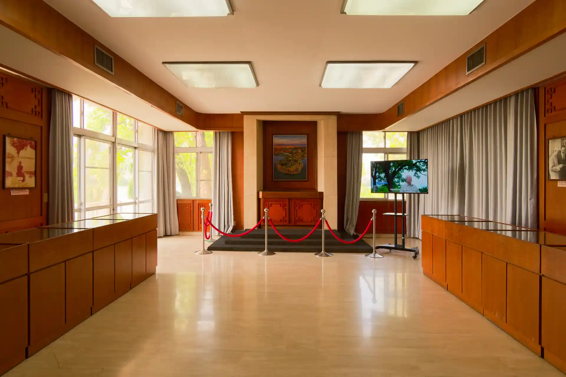 A formal room with a vinyl floor and inbuilt wooden cupboards along both sides. At the far end of the room, there is a roped off area around a painting on the wall. There is also a TV on a stand, playing a video.