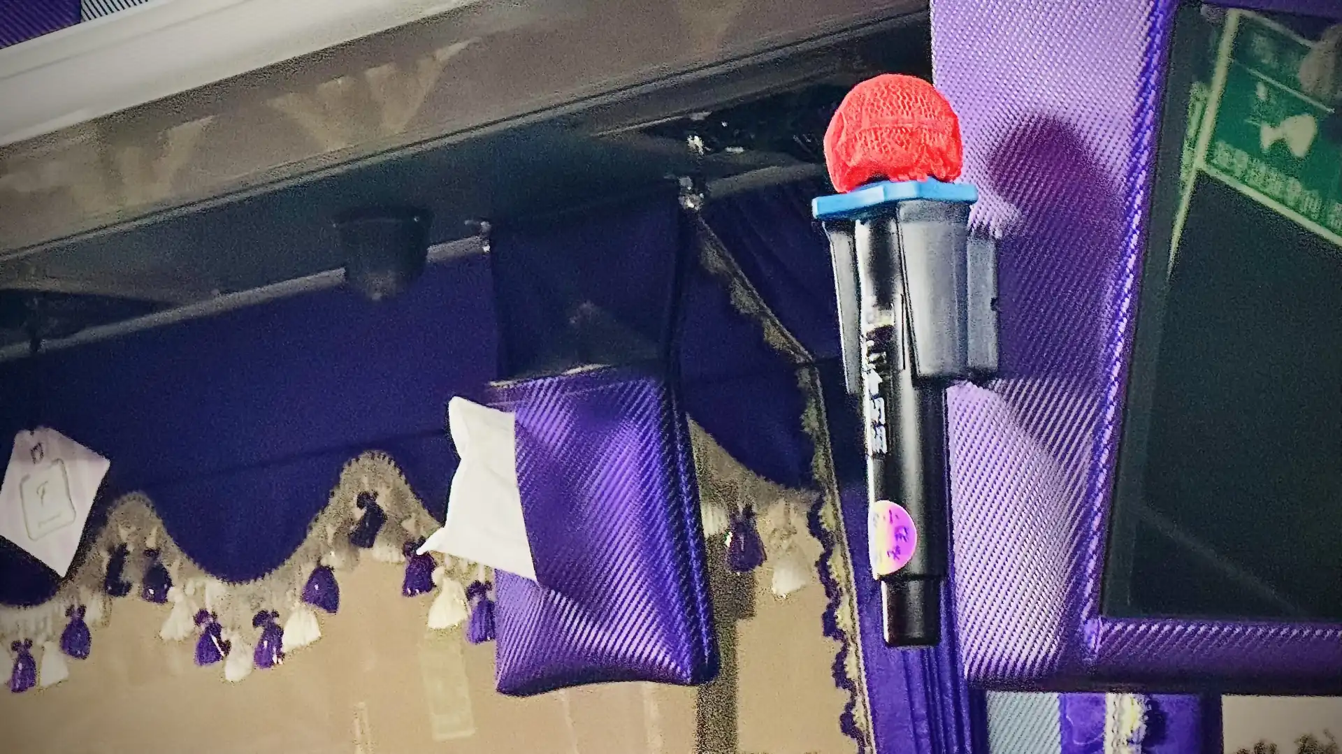 Bus interior close-up of a karaoke microphone mounted to a ceiling-mounted TV screen embedded in a bright purple case, with a matching bright purple tissue holder hanging in front of window valances.