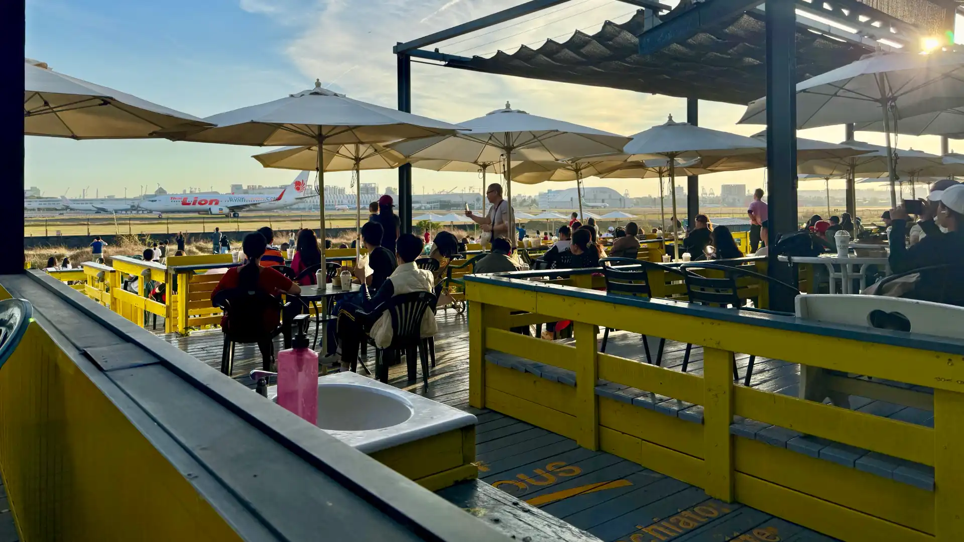 A daytime view of the Old Daddy Farm outdoor cafe. There are dozens of people sitting at tables under sun umbrellas, and looking out towards the airport. A jet is moving down the runway, just beyond the cafe’s boundary fence.