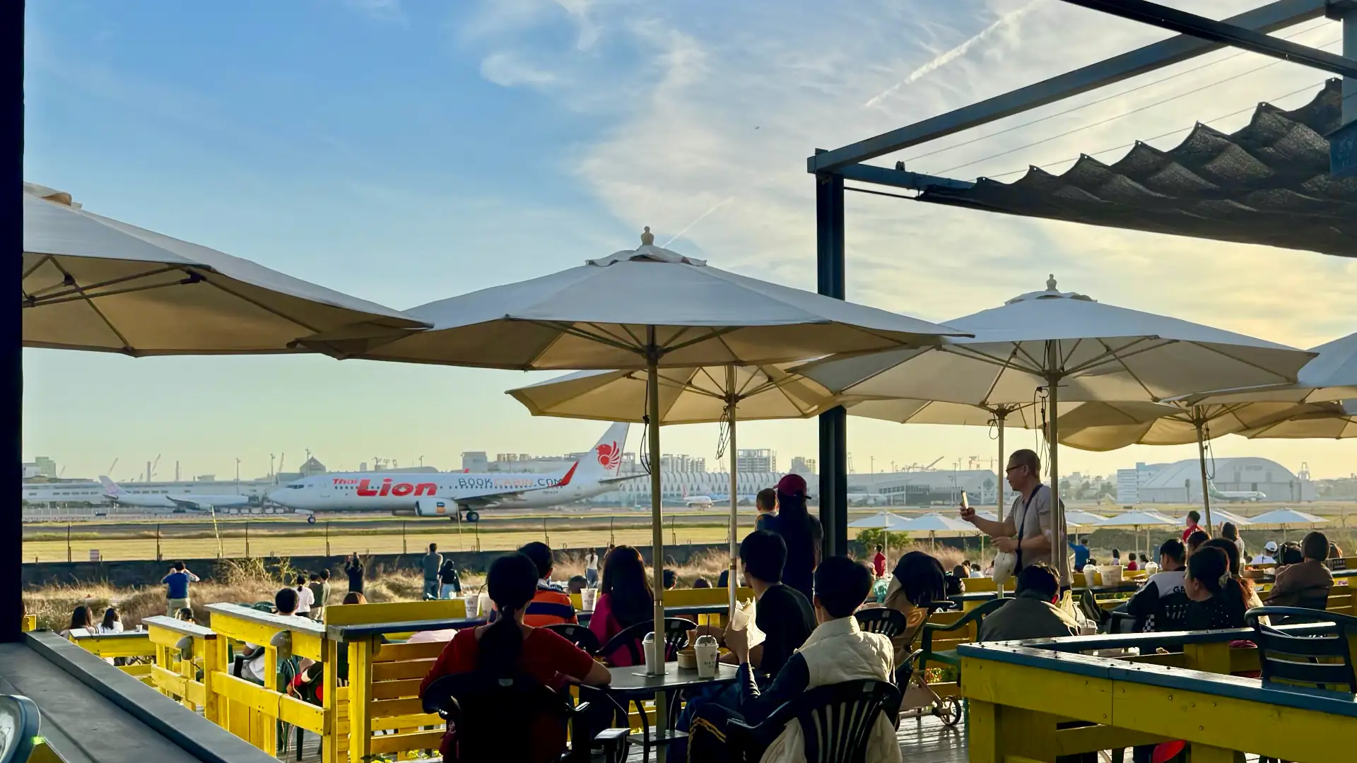 A daytime view of the Old Daddy Farm outdoor cafe. There are dozens of people sitting at tables under sun umbrellas, and looking out towards the airport. A jet is moving down the runway, just beyond the cafe’s boundary fence.