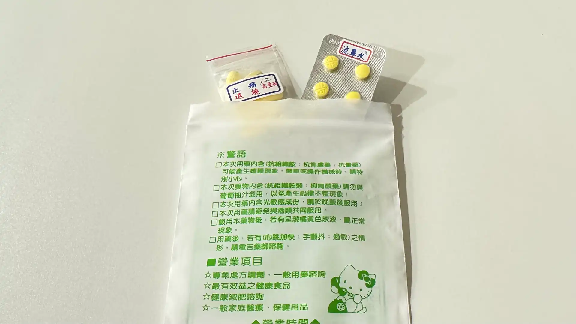 A pack of medicines from a pharmacy in Taiwan. Two different types of pills (one in a blister pack and one in a small plastic bag) are poking out from the top. The bag is covered in printed Chinese. The lower-right side includes a green-and-white cartoon of Hello Kitty using the phone.