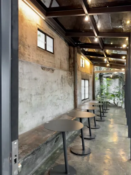 A long narrow brutalist room with concrete bench seating and circular steel tables, looking out through a glass wall.