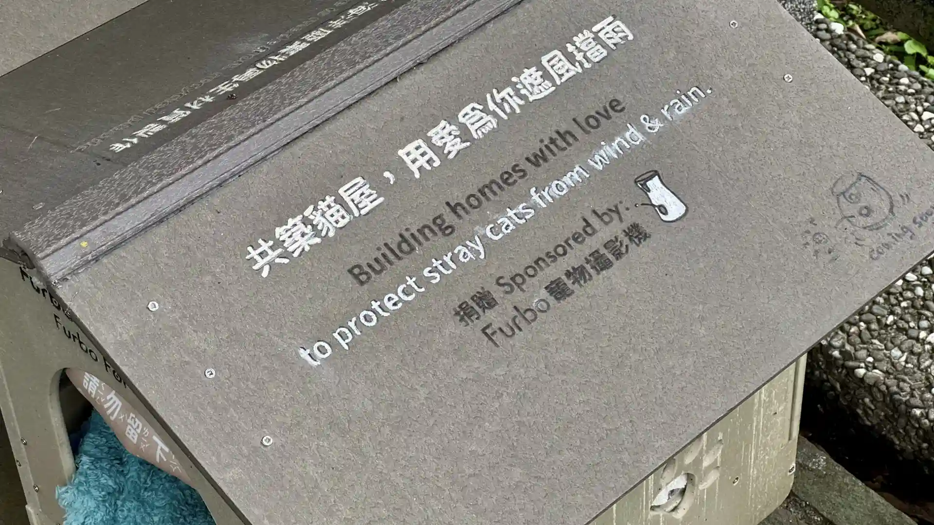 Close-up of a message printed on the roof of a single cat shelter box at Houtong Cat Village. It reads “Building homes with love to protect stray cats from wind and rain. Sponsored by Furbo.”