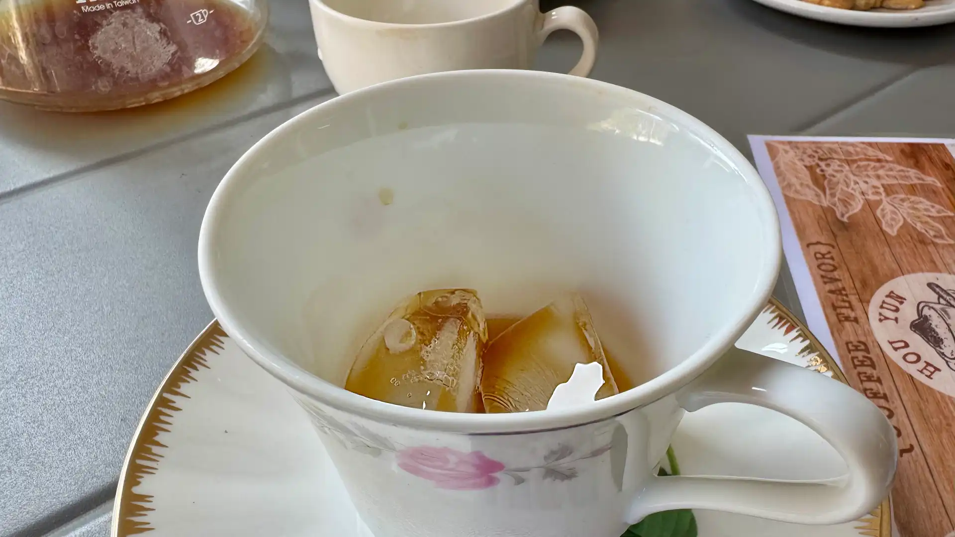 A dainty tea cup containing ice cubes and black coffee.