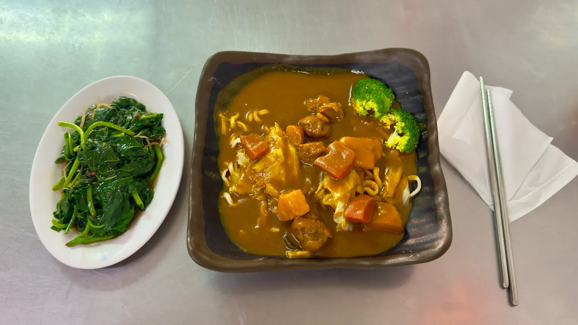 A large bowl of vegan curry ramen next to a smaller side dish of stir-fried sweet potato leaves, and a pair of metal chopsticks.