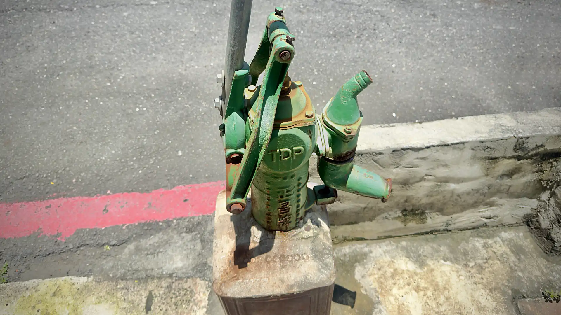 Top-down view of a TDP-branded cast-iron hand-operated water pump, mounted on a concrete base immediately next to a quiet urban street in Central Kaohsiung, Taiwan.