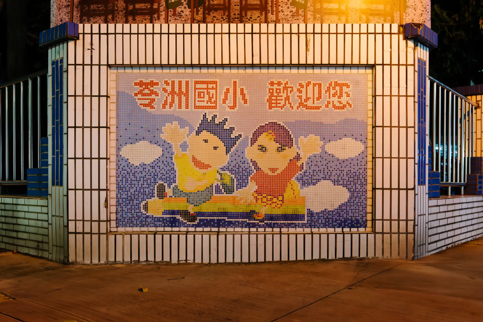 A large mosaic on the tiled fence of a Taiwanese elementary school. It shows two happy children waving, as they ride a giant pencil.