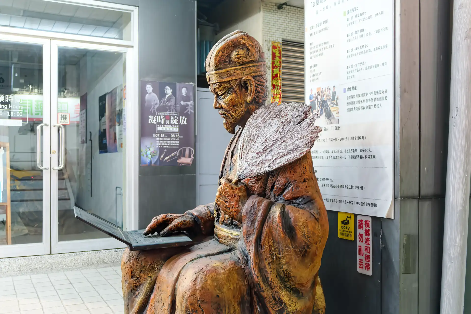 A life-size sculpture of a man in traditional Chinese clothing fanning himself with his left hand and typing on a laptop with his right hand.