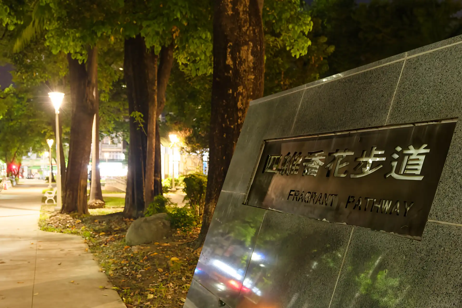 A granite and steel sign that includes Chinese characters and the English name “FRAGRANT PATHWAY”. Behind the sign is a public toilet block in the park.