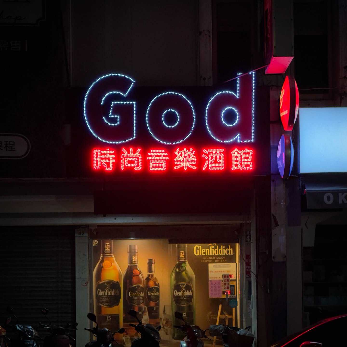 A large sans-serif illuminated sign that reads “God”, covering the second floor of a building that has floor-to-ceiling whisky advertisements in the first floor window.