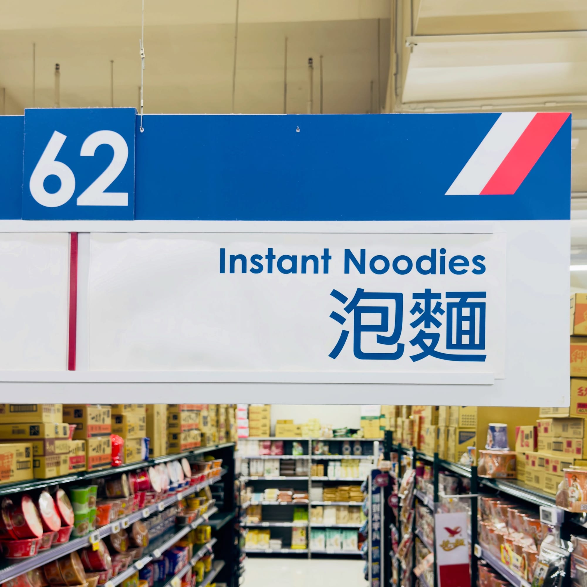 A hanging aisle marker at a Prosperity Plaza supermarket in Taiwan. It is aisle number 62, labeled “Instant Noodies”.
