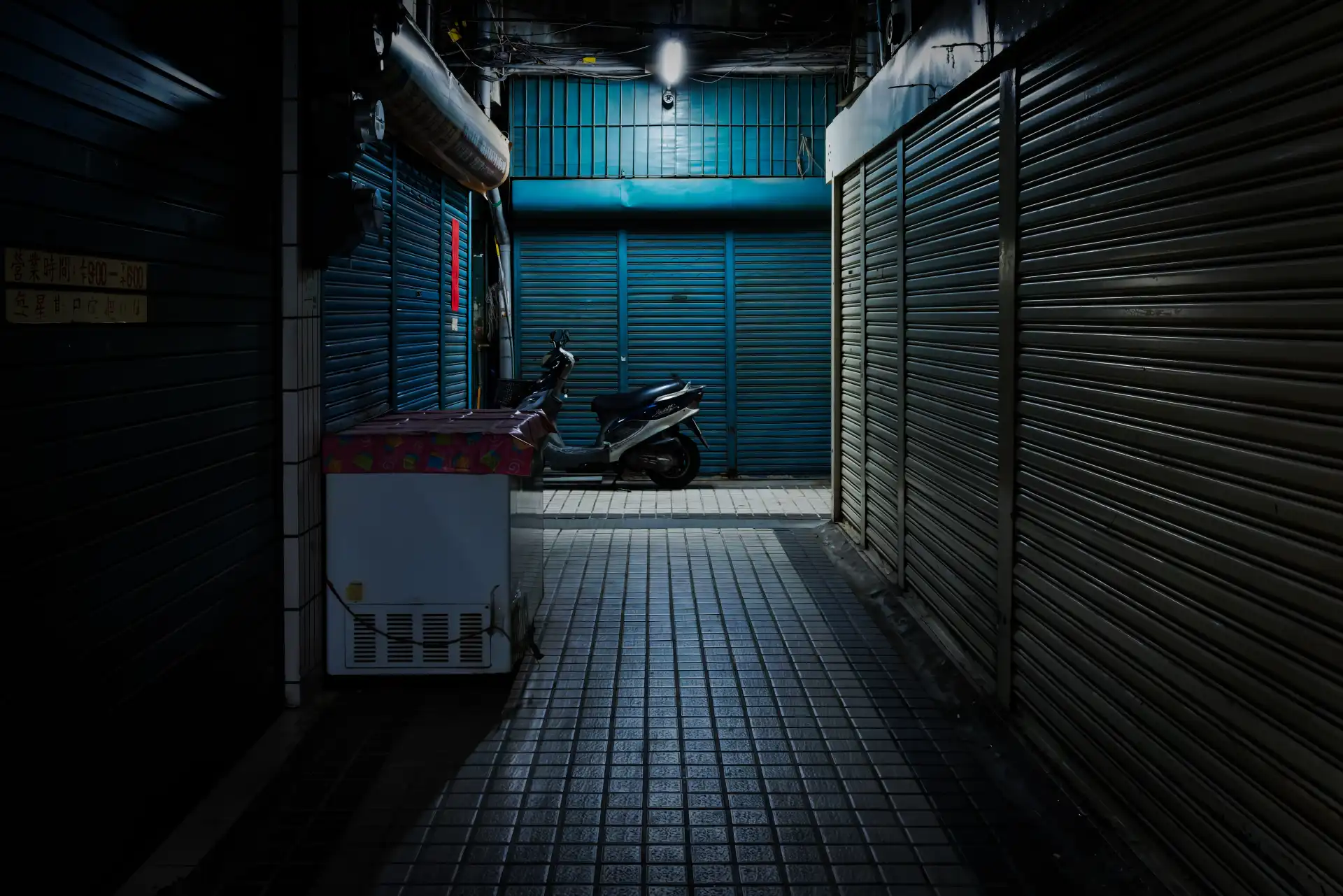 Closed metal roller doors either side of a narrow, dark, lane inside a deserted wet market at night.