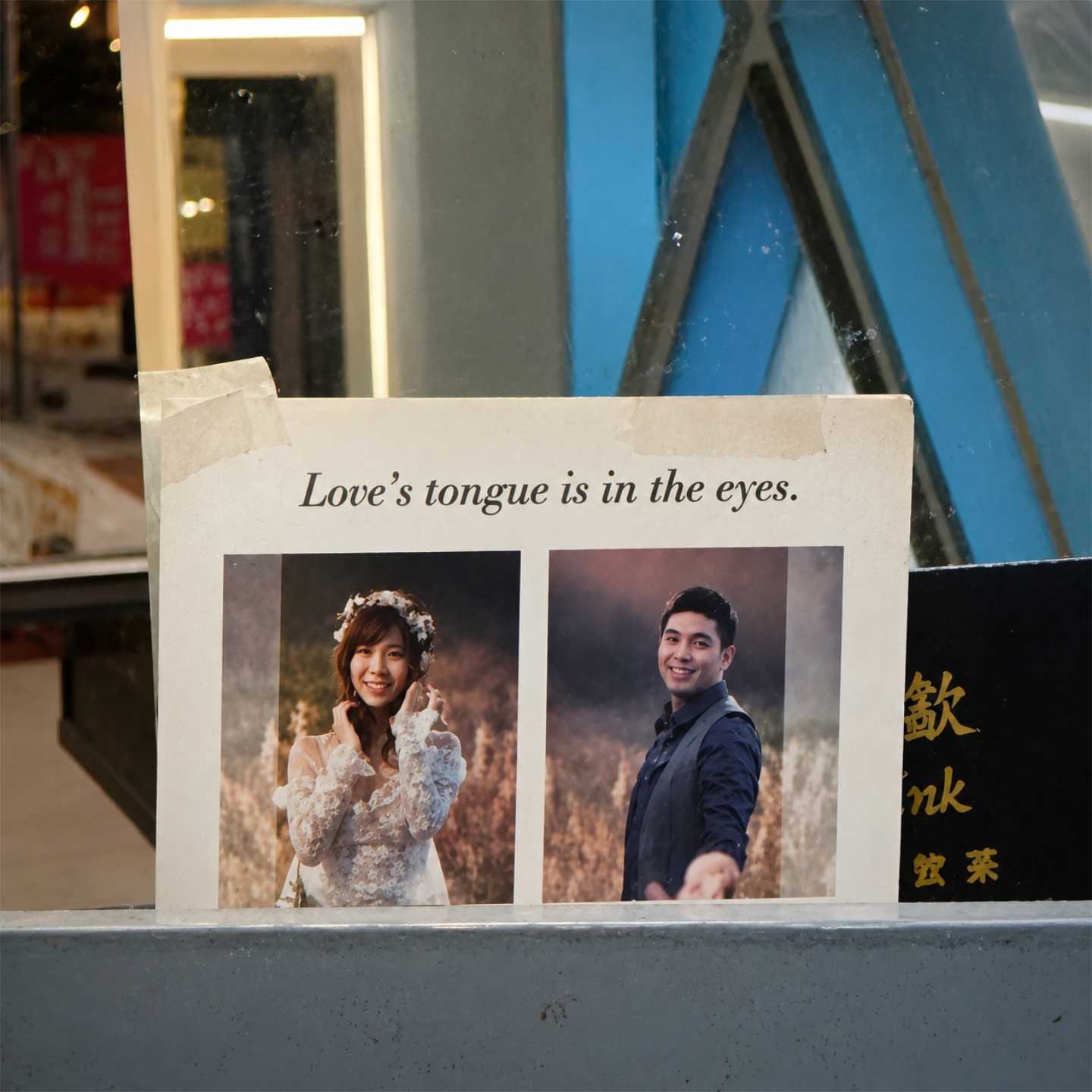A postcard taped to a mirror at a traditional barbershop in Tainan City, Taiwan. It comprises photos of a man and woman standing in a field, with the heading “Love’s tongue is in the eyes”.