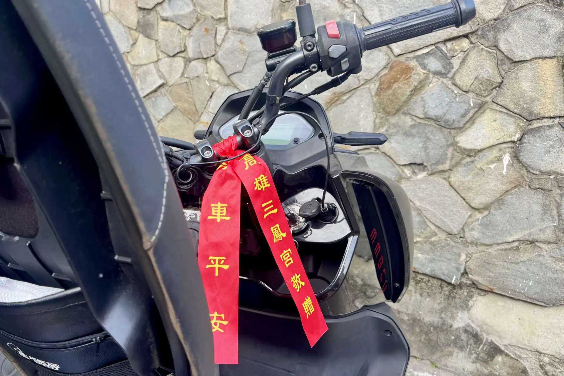 A ribbon-style religious talisman tied to scooter handlebars in Taiwan, for scooter safety. The ribbons are red with gold Traditional Chinese characters on them.