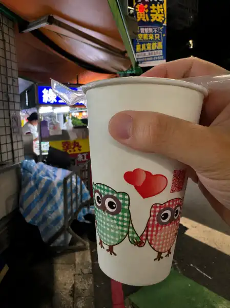 Close-up of a hand holding a large paper cup outside a night market stall.