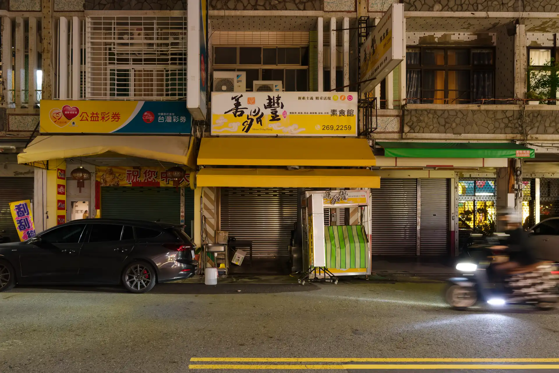 A metal roller door covers the street frontage of a closed vegetarian restaurant in Kaohsiung, Taiwan. The long exposure has captured a blurred person on a scooter riding past.
