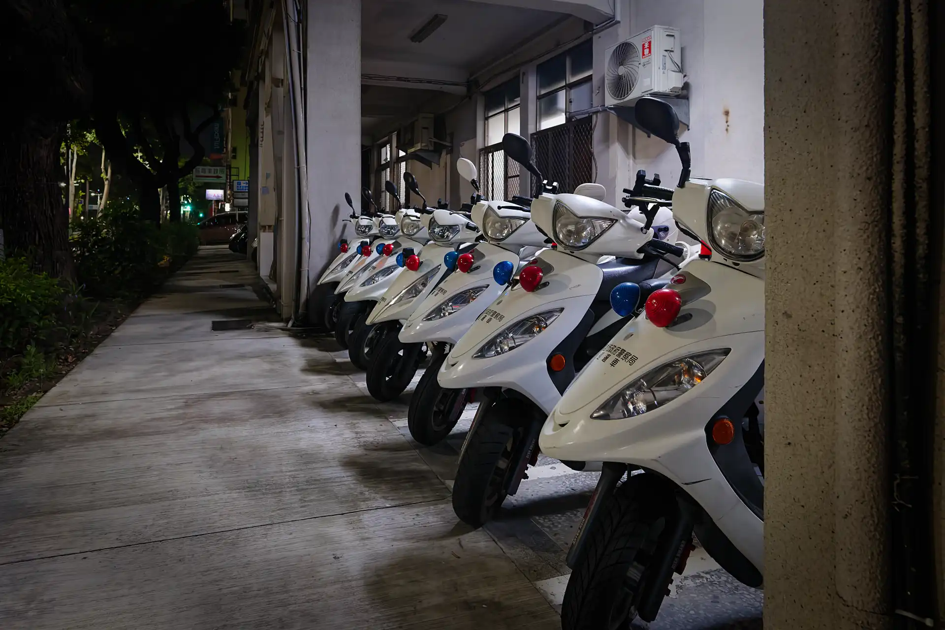 A line-up of seven white 125cc police scooters, with bulbous blue and red lights mounted on the front, parked on the sidewalk outside Chenggong Road Police Station in Kaohsiung, Taiwan.