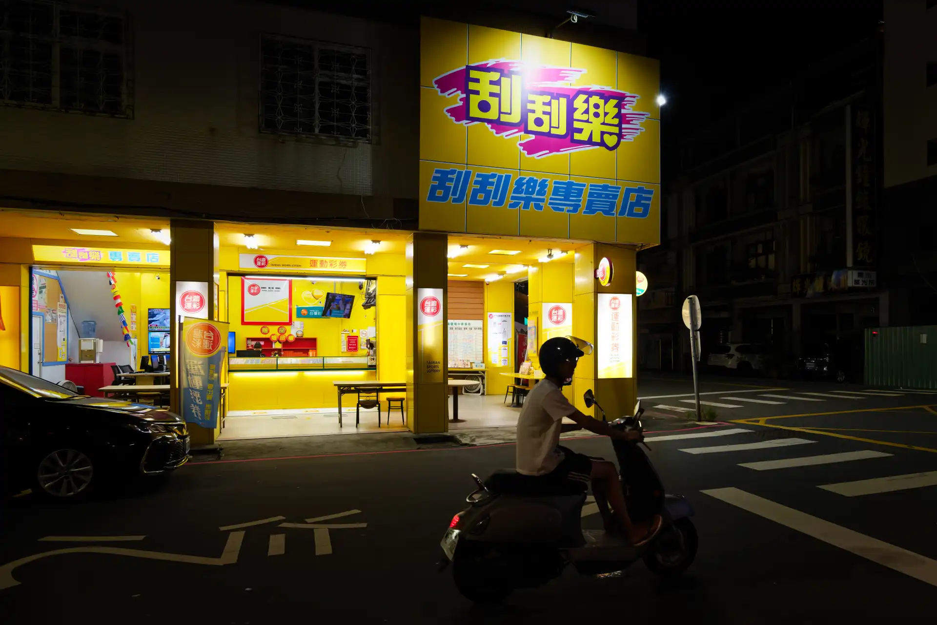 A man on a scooter riding past a branch of the Taiwan Sports Lottery at night.