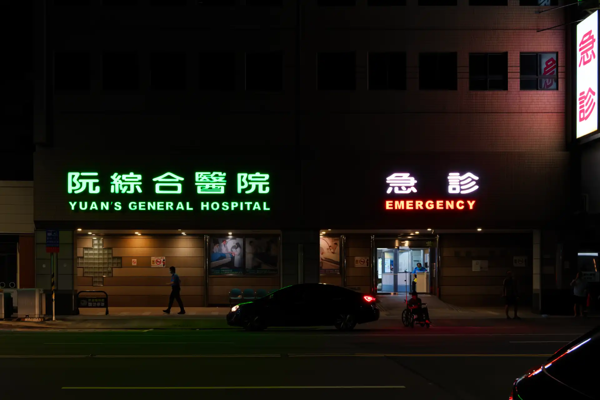 Yuan’s General Hospital emergency entrance at night. A car is parked on the street outside. Its brake lights are illuminating a man sitting in a wheelchair outside the hospital entrance.