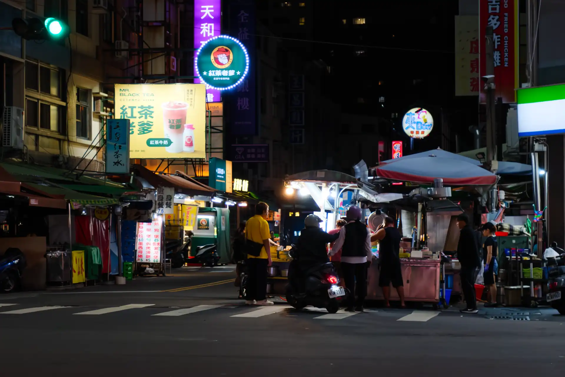 People queueing at a night market stall. One of the people in the queue is sitting on a scooter.