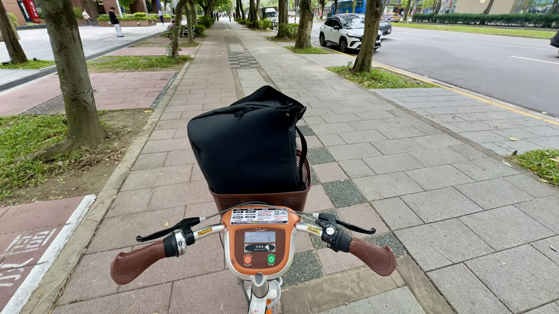 Point-of-view shot of a satchel bag in the front basket of an electric YouBike, riding on a paved sidewalk in Taipei.