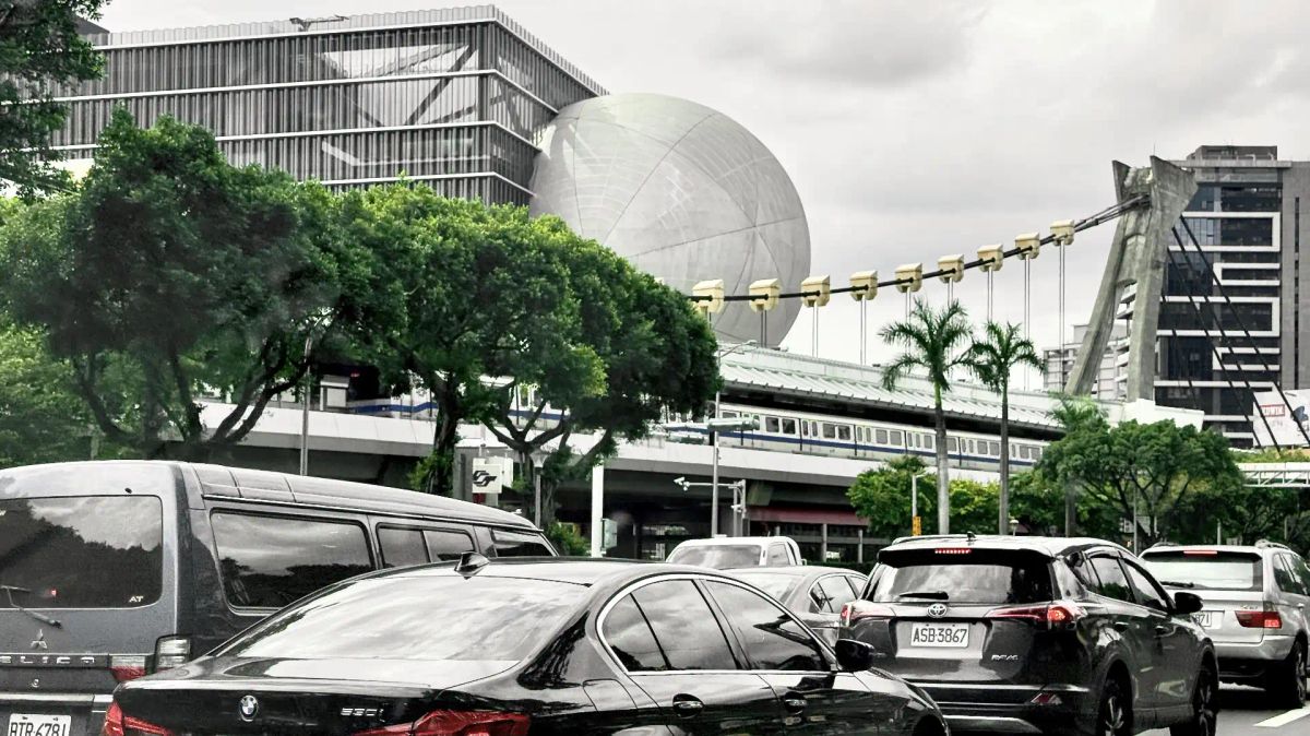 A train stopped at an elevated metro station, above a busy multi-lane road. The avant garde Taipei Performing Arts Center is visible behind the station.