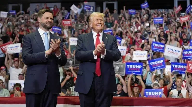 JD Vance and Donald Trump applaud together at a campaign rally with supporters holding Trump-Vance signs
