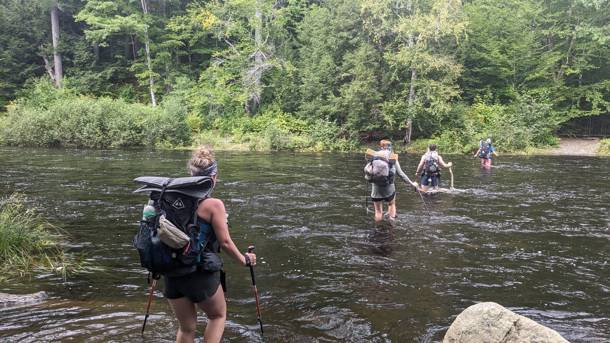 Thru-hikers wading across a broad river in the 100-Mile Wilderness with backpacks held above the water.