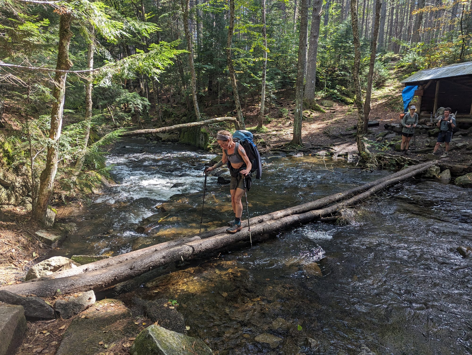 Backpacker balancing across a fallen log over a forest stream in Maine's 100-Mile Wilderness.