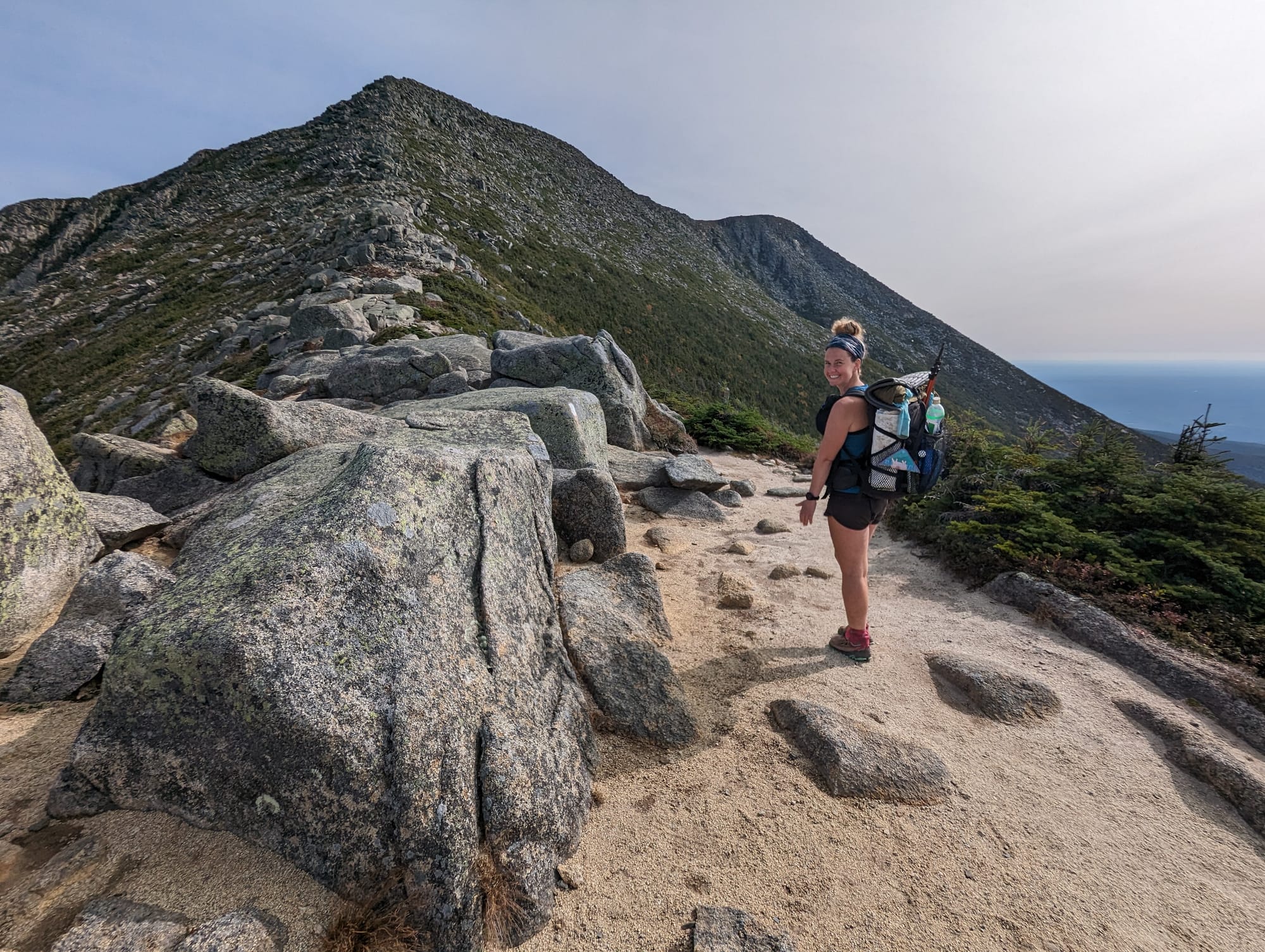 Sunshine hiking across exposed granite with Baxter Peak rising behind her on Mount Katahdin.