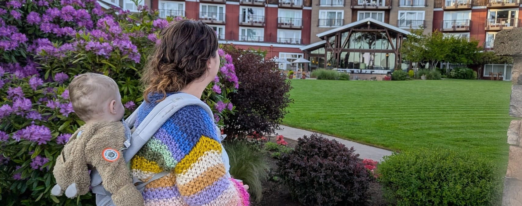 Miranda, in a rainbow sweater, carries her son as the walk around Suquamish: purple rhododendrons and lodge-style building in the background.
