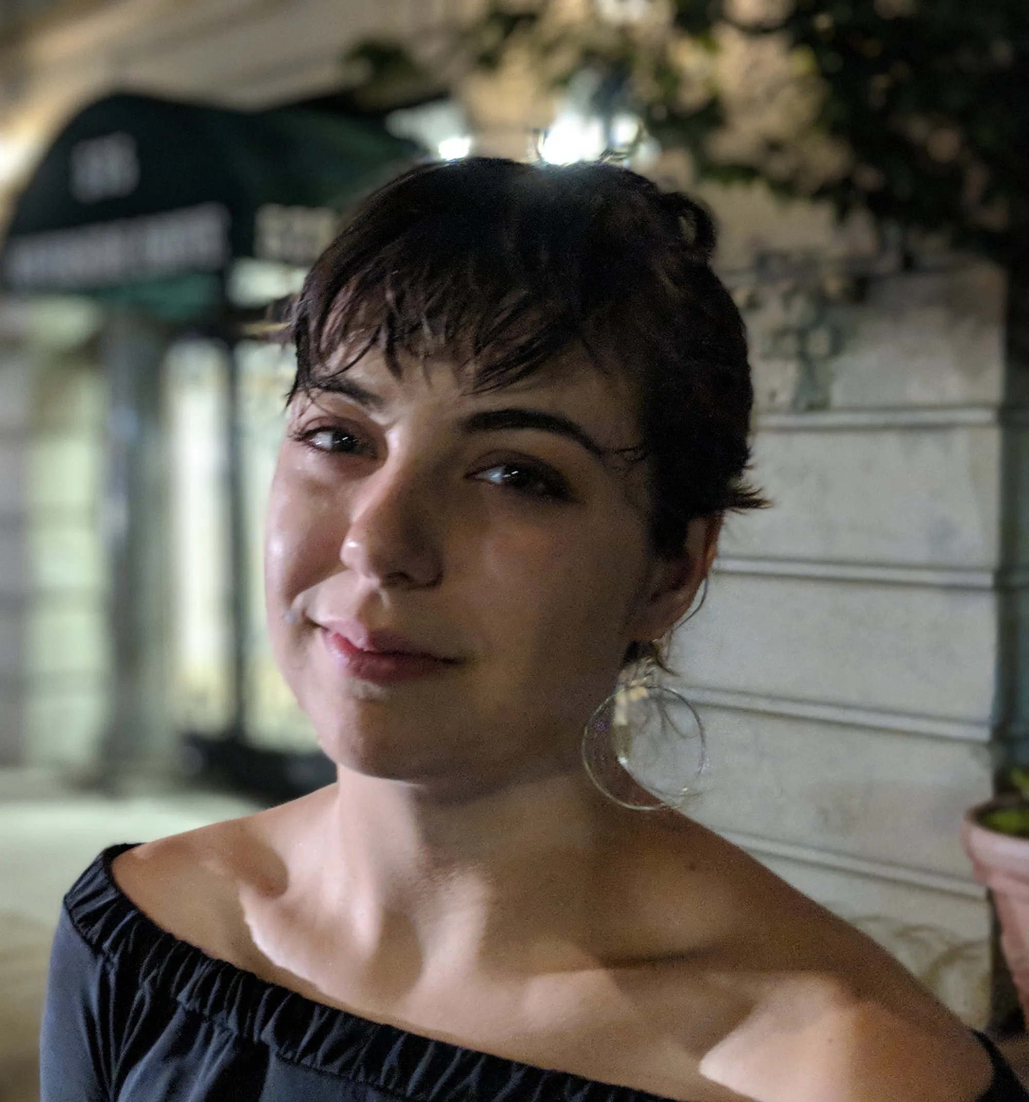 Miranda Sita smiling outdoors at night, wearing an off the shoulder black top and large hoop earrings.