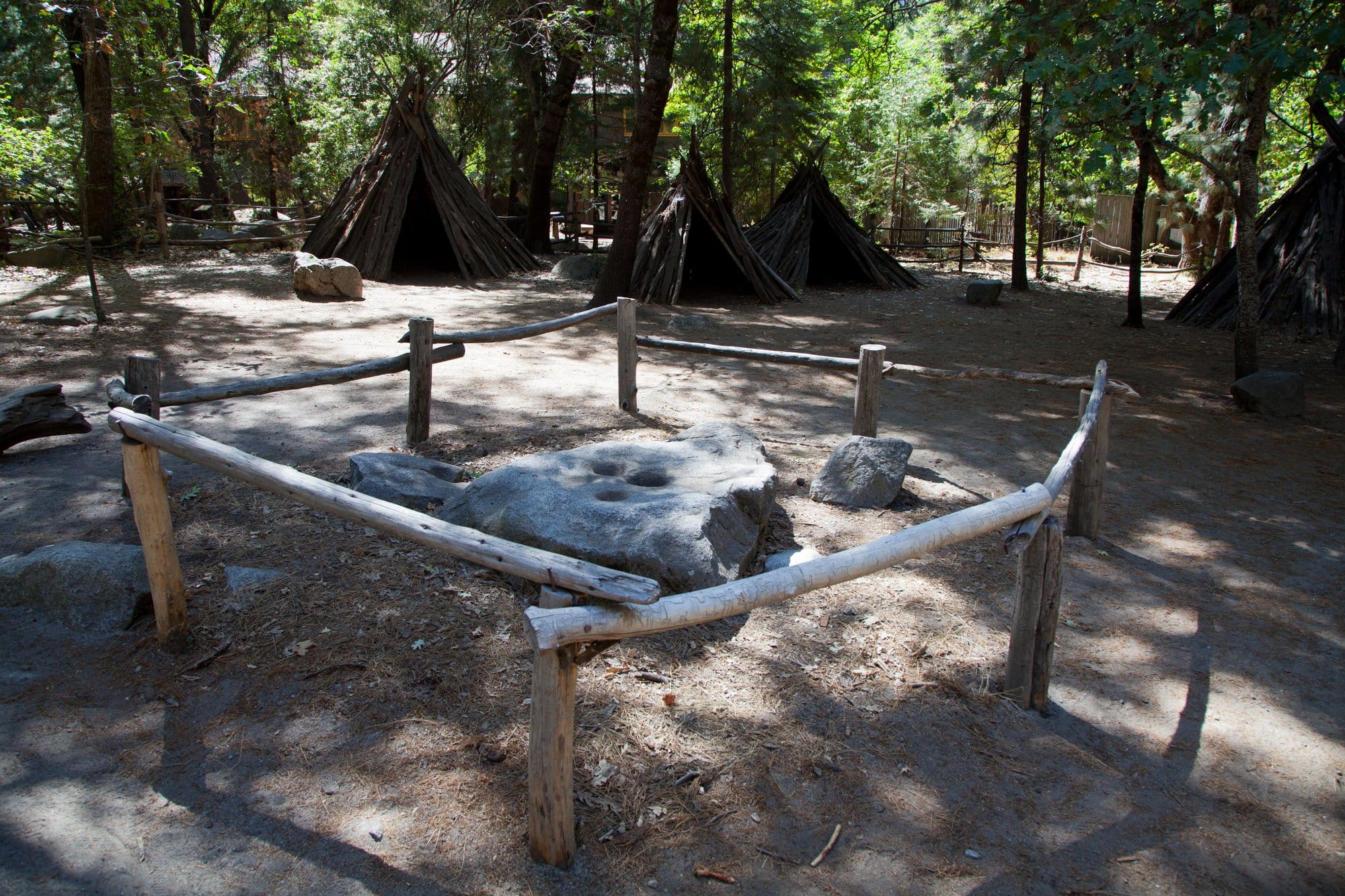 A low granite boulder with several round, shallow grinding holes, inside a rail fence, with bark-covered conical shelters among the trees behind.
