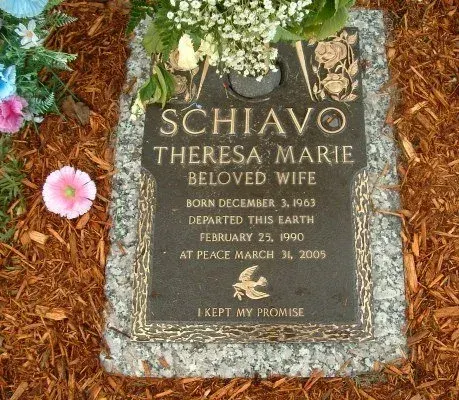 Bronze grave marker reading Schiavo, Theresa Marie, Beloved Wife, with flowers and wood mulch around it.