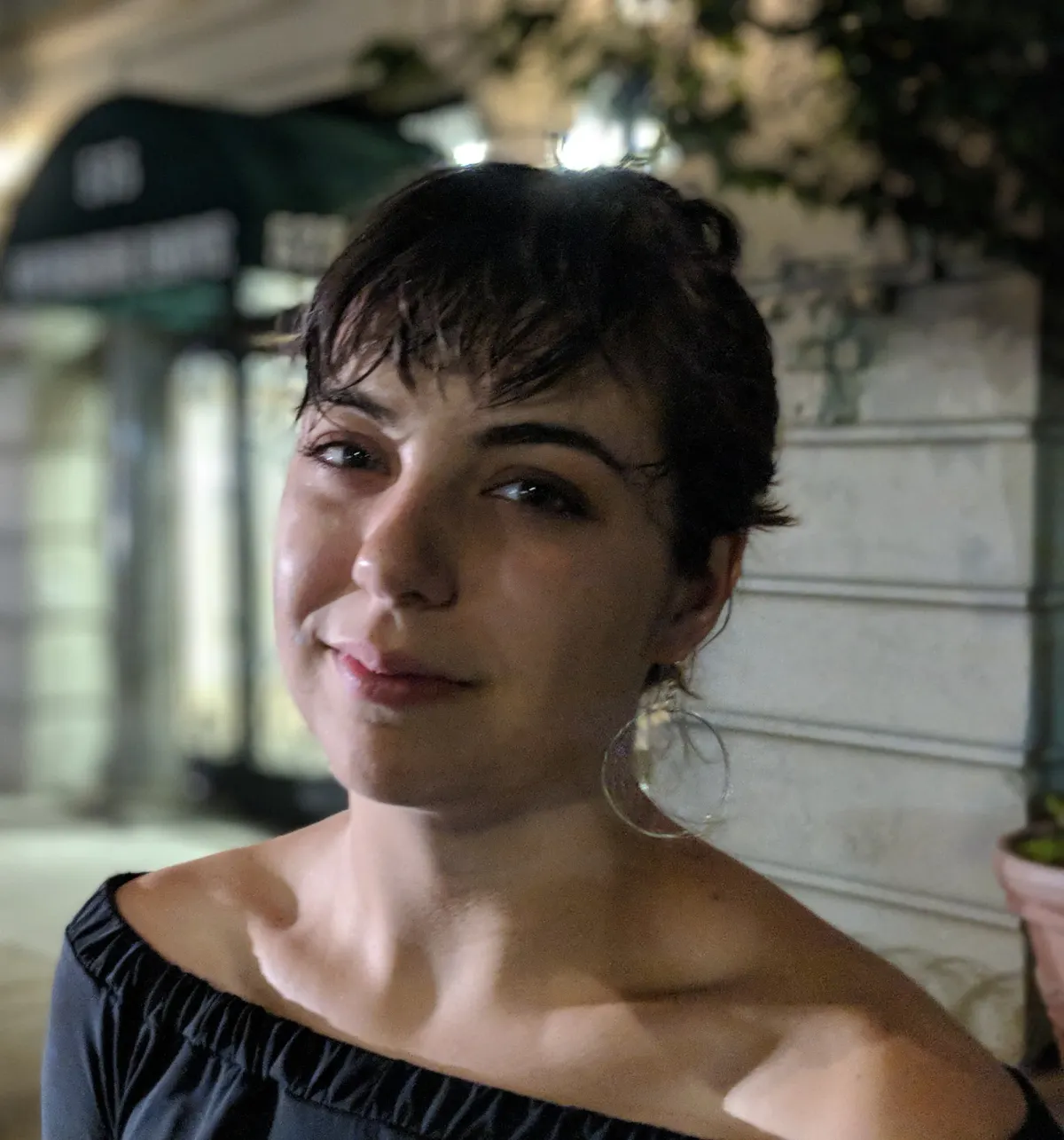 Miranda Sita smiling outdoors at night, wearing an off the shoulder black top and large hoop earrings.